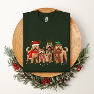 May include: Four golden doodle dogs wearing festive Christmas attire, including Santa hats, reindeer antlers, and elf hats, with a string of Christmas lights in front of them. The dogs are standing on a green background.
