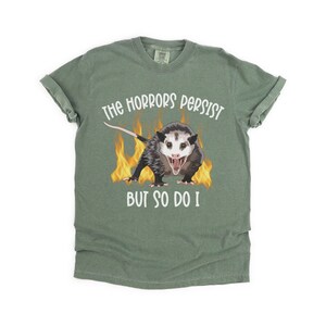 K&ouml;nnte beinhalten: Ein gr&uuml;nes T-Shirt mit einem wei&szlig;en Aufdruck eines Opossums in Flammen mit dem Text "The horrors persist but so do I."