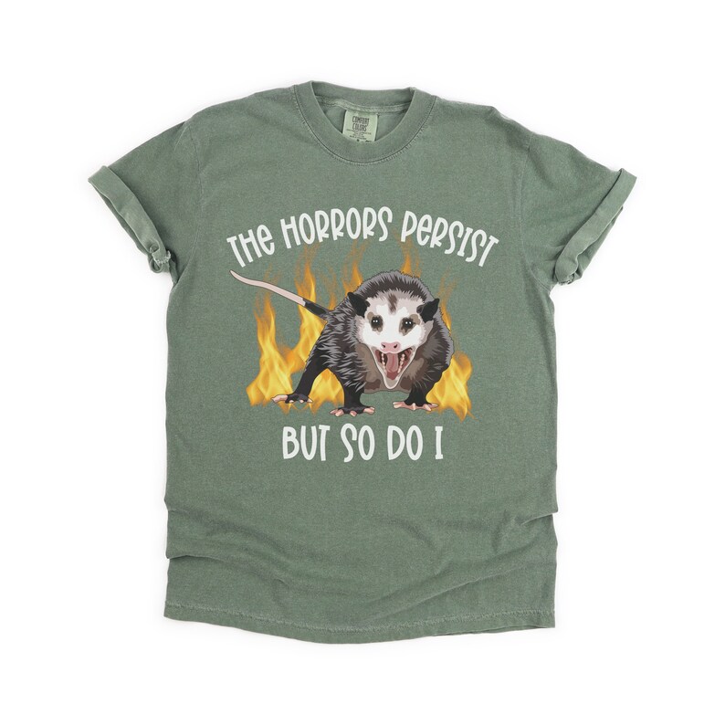 K&ouml;nnte beinhalten: Ein gr&uuml;nes T-Shirt mit einem wei&szlig;en Aufdruck eines Opossums in Flammen mit dem Text "The horrors persist but so do I."