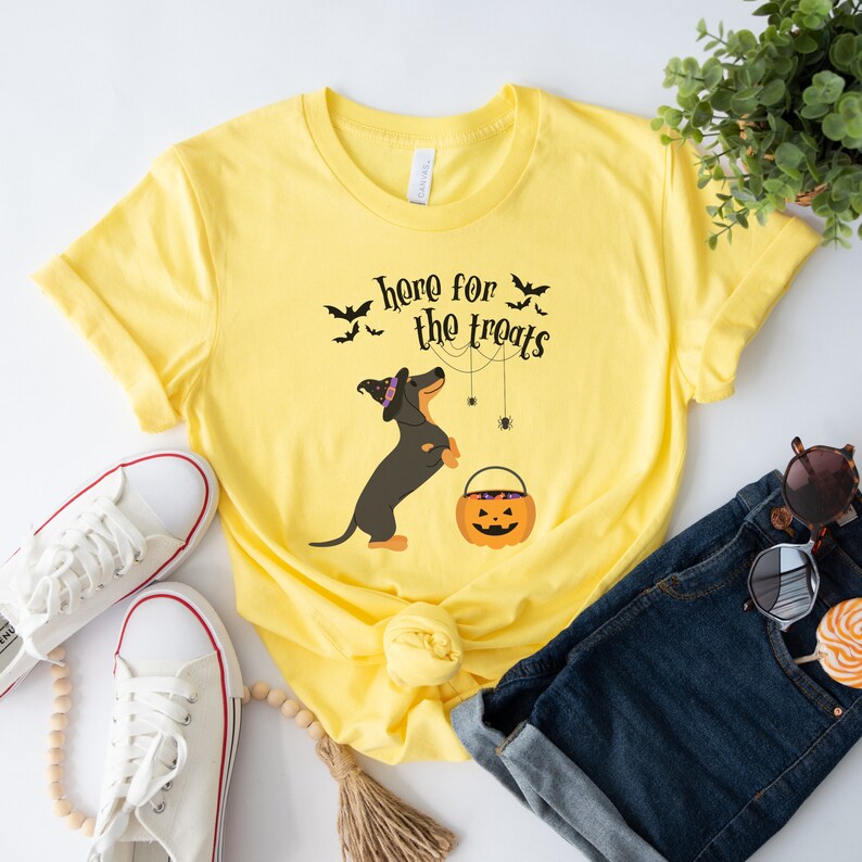 K&ouml;nnte beinhalten: Gelbes T-Shirt mit einem schwarzen Dackel, der einen Hexenhut tr&auml;gt und einen K&uuml;rbis-Eimer h&auml;lt. Der Text "Here for the treats" ist auf dem T-Shirt aufgedruckt.