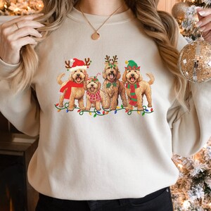 May include: A cream-colored sweatshirt with a Christmas-themed design featuring four doodle dogs wearing festive hats and scarves. The dogs are standing in front of a string of Christmas lights.