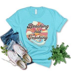 May include: A turquoise blue t-shirt with a retro-style graphic design. The graphic features a sun, clouds, and waves with the text "Beaching not Teaching".