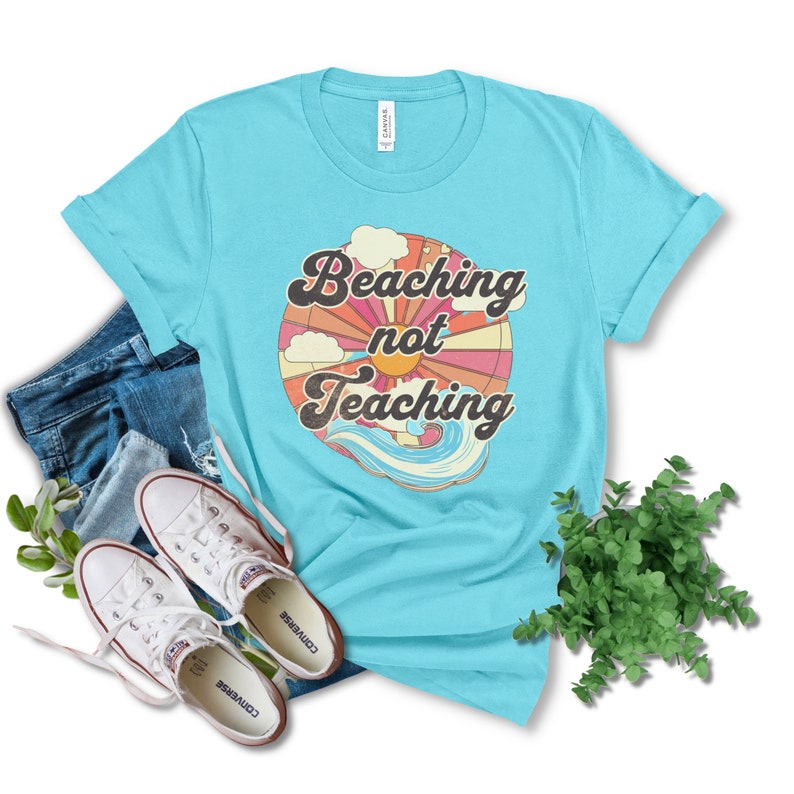 May include: A turquoise blue t-shirt with a retro-style graphic design. The graphic features a sun, clouds, and waves with the text "Beaching not Teaching".