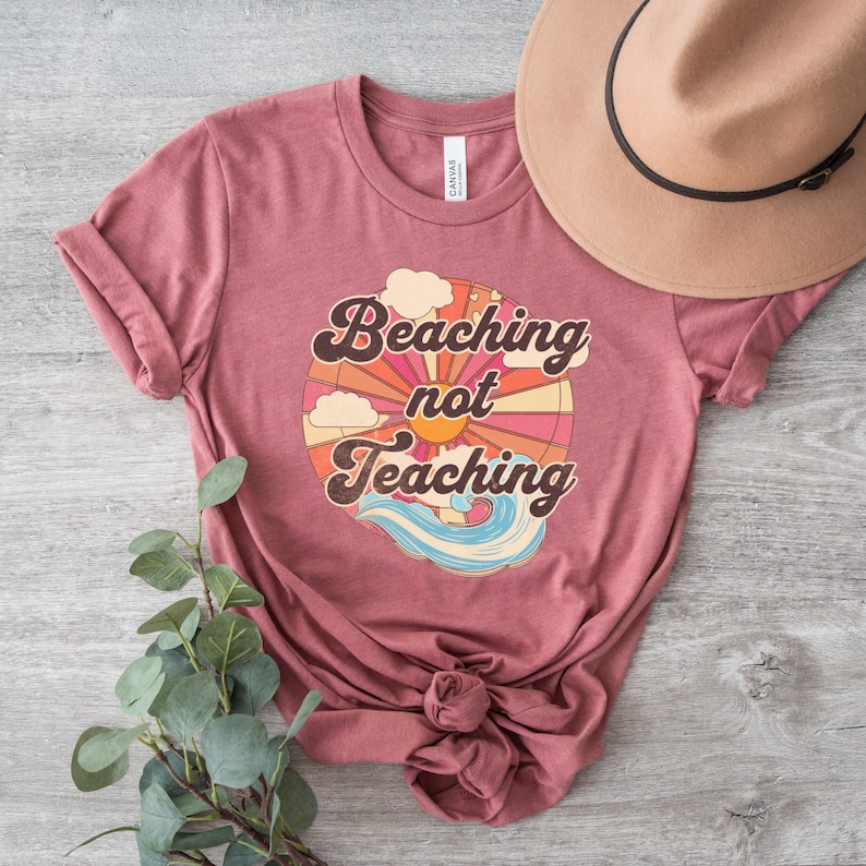 May include: A maroon t-shirt with a retro-style graphic that says "Beaching not Teaching" with a sun, clouds, and waves.
