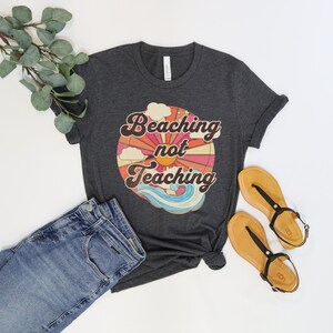 May include: A dark heather grey t-shirt with a retro-style graphic that says "Beaching not Teaching" with a sun, clouds, and waves.