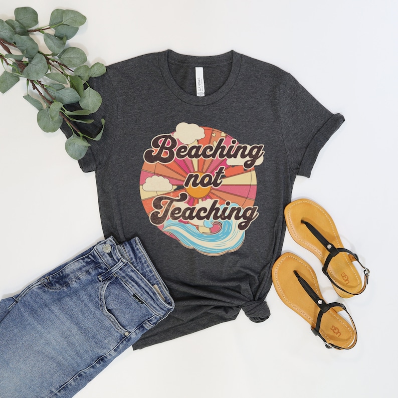 May include: A dark heather grey t-shirt with a retro-style graphic that says "Beaching not Teaching" with a sun, clouds, and waves.