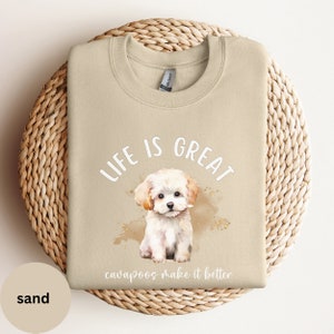 K&ouml;nnte beinhalten: Ein beiger Pullover mit dem Text "Life is Great Cavapoos make it better" und einer Aquarell-Illustration eines wei&szlig;en Cavapoo-Welpen.