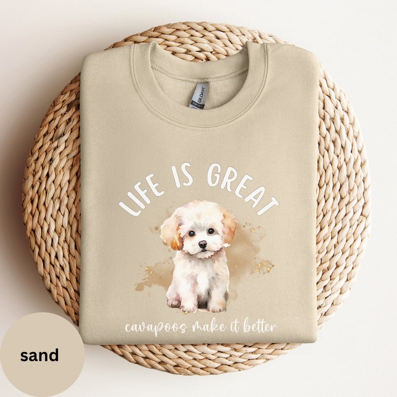 K&ouml;nnte beinhalten: Ein beiger Pullover mit dem Text "Life is Great Cavapoos make it better" und einer Aquarell-Illustration eines wei&szlig;en Cavapoo-Welpen.