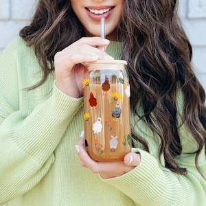 May include: A clear glass reusable drinking bottle with a bamboo lid and a straw. The bottle is decorated with a pattern of colorful chickens and flowers.