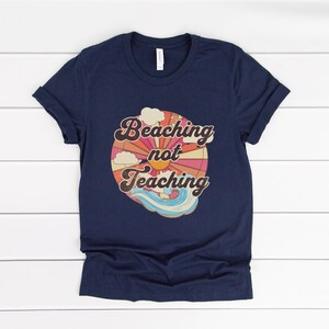 May include: Navy blue t-shirt with a retro-style graphic of a sun, clouds, and waves. The graphic says "Beaching not Teaching".
