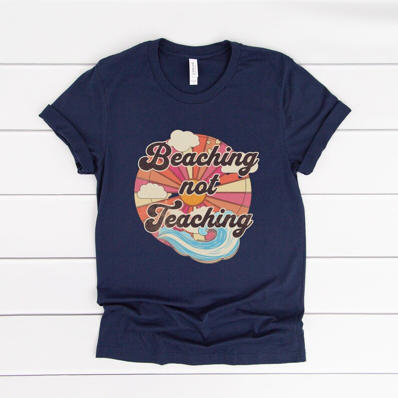 May include: Navy blue t-shirt with a retro-style graphic of a sun, clouds, and waves. The graphic says "Beaching not Teaching".