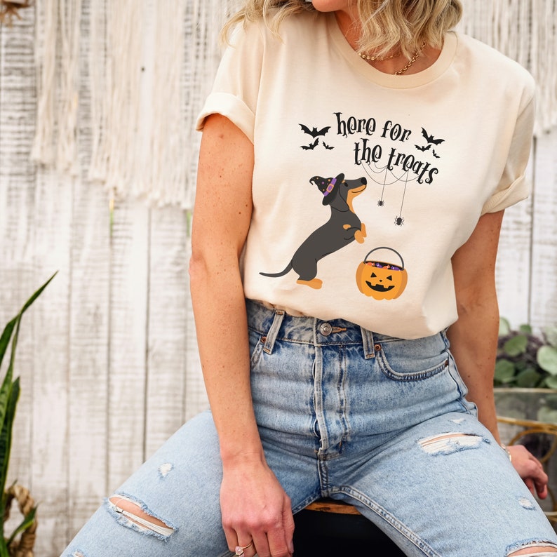 K&ouml;nnte beinhalten: Ein cremefarbenes T-Shirt mit einem schwarzen Dackel, der einen Hexenhut tr&auml;gt und einen K&uuml;rbis-Eimer h&auml;lt. Der Text auf dem Shirt lautet "here for the treats" mit Flederm&auml;usen und Spinnweben, die den Hund umgeben.