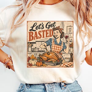 Pode incluir: Uma t-shirt cor de creme com um gráfico de estilo retro de uma pessoa com avental a regar um peru assado numa cozinha. O texto diz Let's Get BASTED e Good Food Good Company. A ilustração inclui abóboras, folhas de outono e vinho.