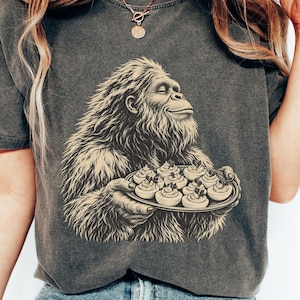 May include: A charcoal gray short-sleeve t-shirt featuring a graphic print of a hairy, ape-like creature holding a platter of deviled eggs. The creature has a content expression with eyes closed. The shirt has a vintage, washed-out appearance.