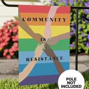 May include: A garden flag with a rainbow design and the words "COMMUNITY IS RESISTANCE." The flag features illustrations of hands of various skin tones joined together. The flag is displayed on a black metal garden stake.