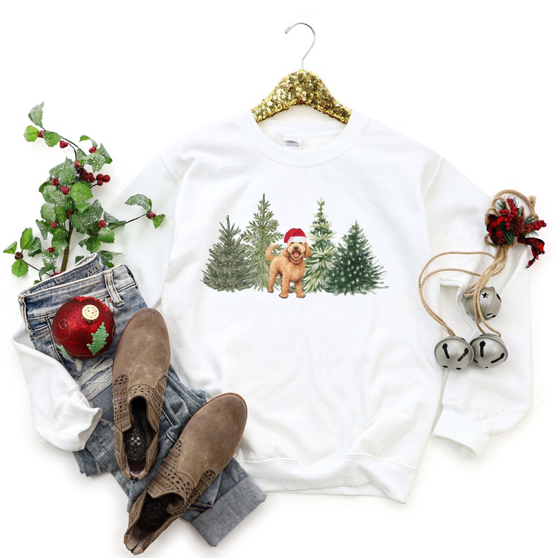 May include: White Christmas sweatshirt with a golden doodle dog wearing a Santa hat standing in front of a row of Christmas trees.