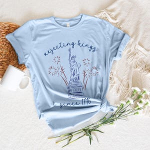 May include: Pale blue t-shirt with a graphic of the Statue of Liberty, fireworks, and the text "rejecting kings since 1776". The shirt is knotted at the waist and is on a neutral background.