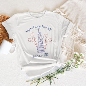 May include: White t-shirt with a blue and red graphic of the Statue of Liberty, fireworks, and the text "rejecting kings since 1776". The shirt is knotted at the waist.