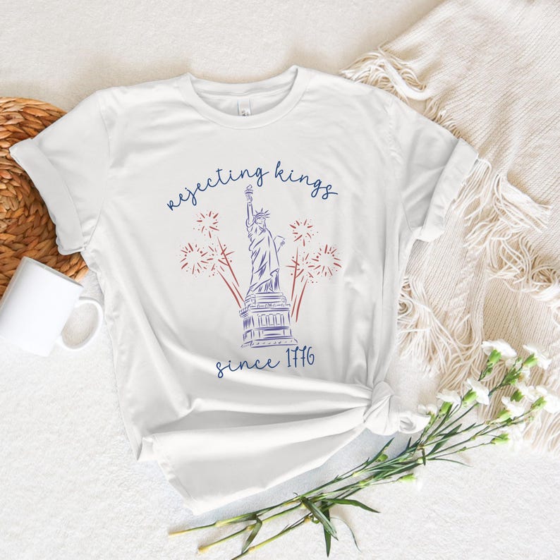 May include: White t-shirt with a blue and red graphic of the Statue of Liberty, fireworks, and the text "rejecting kings since 1776". The shirt is knotted at the waist.