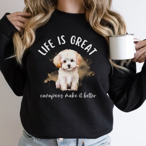 K&ouml;nnte beinhalten: Schwarzer Pullover mit einer wei&szlig;en Aquarell-Illustration eines kleinen wei&szlig;en Hundes. Der Text "Life is Great Cavapoos make it better" ist auf dem Pullover aufgedruckt.