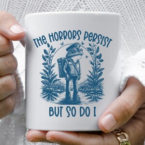 May include: White ceramic mug with a blue illustration of a frog wearing a mail carrier uniform and a backpack. The text "The Horrors Persist But So Do I" is printed on the mug.