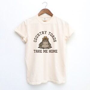 以下が含まれることがあります： クリーム色のTシャツに、カウボーイハットをかぶった茶色のカエルの漫画が描かれています。Tシャツには「COUNTRY TOADS TAKE ME HOME」という文字が印刷されています。