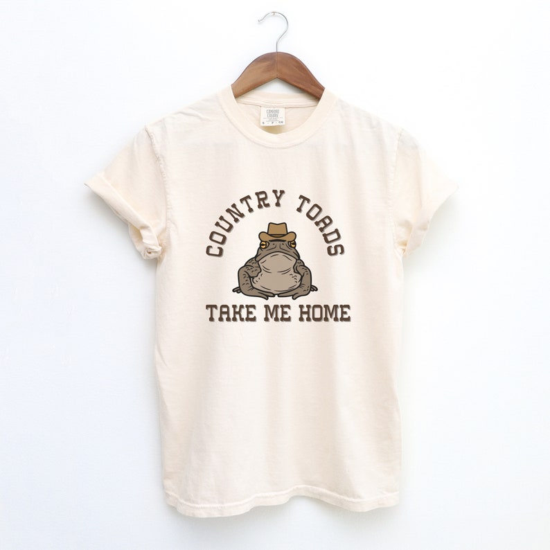 以下が含まれることがあります： クリーム色のTシャツに、カウボーイハットをかぶった茶色のカエルの漫画が描かれています。Tシャツには「COUNTRY TOADS TAKE ME HOME」という文字が印刷されています。