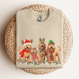 May include: A beige sweatshirt with a Christmas-themed design featuring four doodle dogs wearing festive hats and a string of lights. The dogs are wearing Santa hats, reindeer antlers, elf hats, and a candy cane.