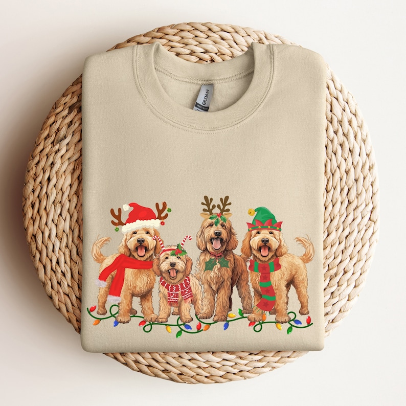 May include: A beige sweatshirt with a Christmas-themed design featuring four doodle dogs wearing festive hats and a string of lights. The dogs are wearing Santa hats, reindeer antlers, elf hats, and a candy cane.