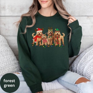 May include: A forest green sweatshirt with a Christmas-themed design featuring four doodle dogs wearing festive hats and antlers. The dogs are standing in front of a string of Christmas lights.