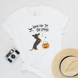 K&ouml;nnte beinhalten: Wei&szlig;es T-Shirt mit einem schwarzen Dackel, der einen Hexenhut tr&auml;gt und einen S&uuml;&szlig;igkeiteneimer h&auml;lt. Der Text "here for the treats" ist auf das Shirt gedruckt.