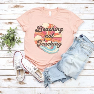 May include: A pink t-shirt with a retro sun graphic and the text "Beaching not Teaching". The shirt is laid out on a white wood background with a pair of white sneakers and blue jeans.