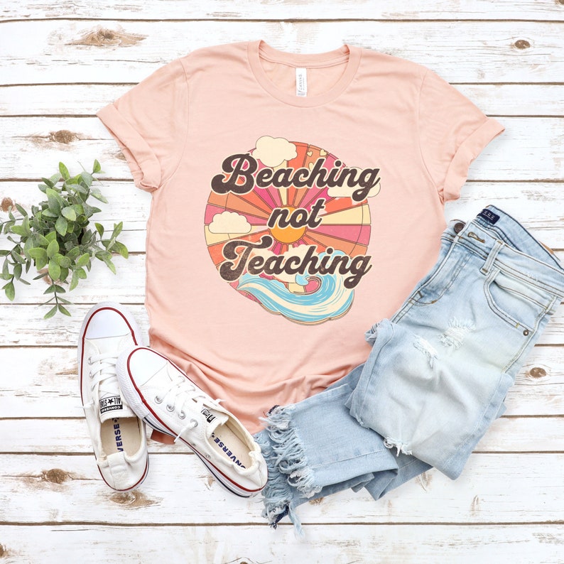 May include: A pink t-shirt with a retro sun graphic and the text "Beaching not Teaching". The shirt is laid out on a white wood background with a pair of white sneakers and blue jeans.
