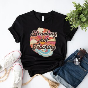 May include: Black t-shirt with a retro-style graphic that says "Beaching not Teaching" with a sun, clouds, and waves.