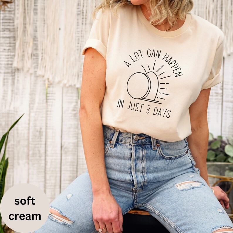 Pode incluir: Uma camiseta cor creme com um gr&aacute;fico preto que diz "A lot can happen in just 3 days". O gr&aacute;fico apresenta um sol estilizado nascendo no horizonte.