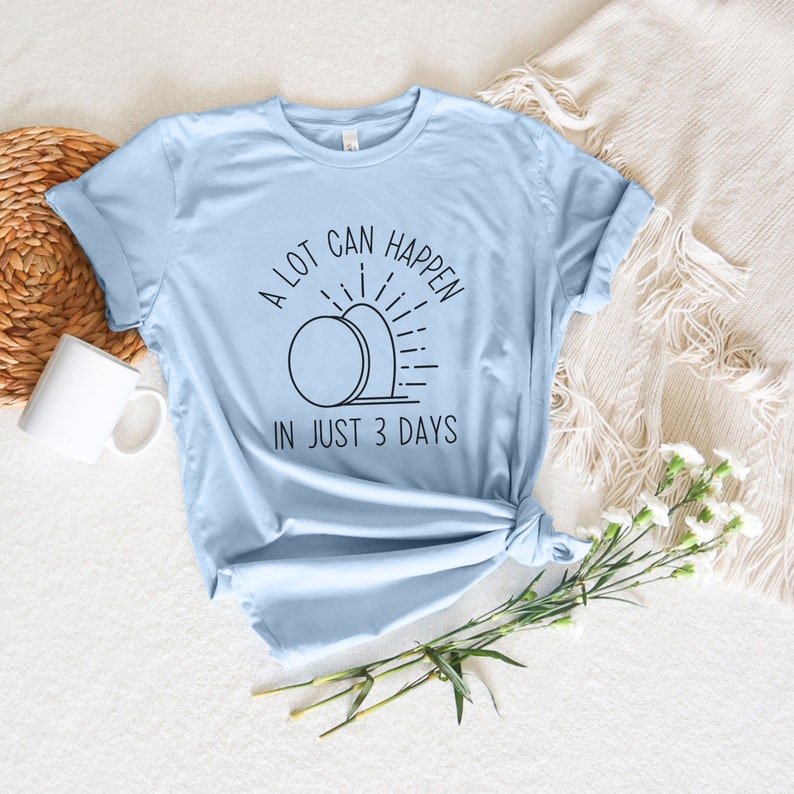 Pode incluir: Uma camiseta azul claro com um design gr&aacute;fico de um nascer do sol sobre uma pedra. O texto "A lot can happen in just 3 days" est&aacute; impresso na camiseta.