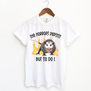 K&ouml;nnte beinhalten: Wei&szlig;es T-Shirt mit einem Aufdruck eines Opossums mit Flammen dahinter. Der Text "The horrors persist but so do I" ist auf das T-Shirt gedruckt.