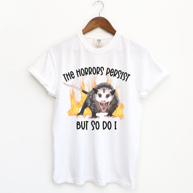 K&ouml;nnte beinhalten: Wei&szlig;es T-Shirt mit einem Aufdruck eines Opossums mit Flammen dahinter. Der Text "The horrors persist but so do I" ist auf das T-Shirt gedruckt.