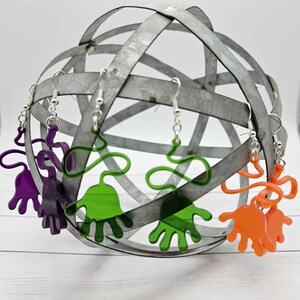Peut inclure: Trois paires de boucles d'oreilles en forme de main sur un pr&eacute;sentoir en m&eacute;tal. Les boucles d'oreilles sont en acrylique transparent et sont disponibles en violet, vert et orange.