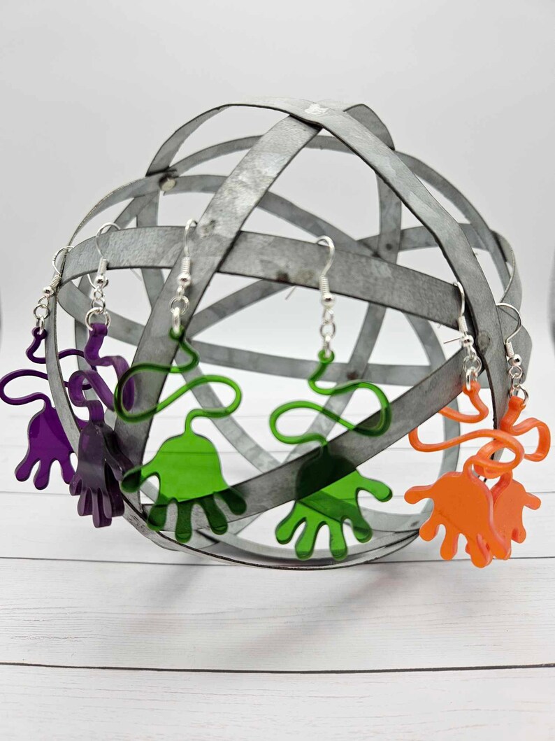 Peut inclure: Trois paires de boucles d'oreilles en forme de main sur un pr&eacute;sentoir en m&eacute;tal. Les boucles d'oreilles sont en acrylique transparent et sont disponibles en violet, vert et orange.