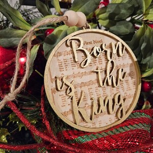 May include: A wooden Christmas ornament with a gold background and the words "Born is the King" in gold lettering. The ornament is decorated with a musical note pattern and the words "The First Noel".