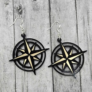 May include: A pair of black and gold compass earrings. The earrings are made of acrylic and feature a gold compass design with black accents.