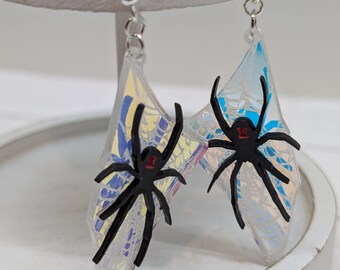 May include: A pair of iridescent spider earrings. Each earring features a clear, wing-shaped design with a black spider attached. The spiders have red accents. The earrings hang from silver-colored hooks.