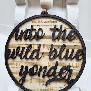 May include: A round wooden ornament with a black border and the words "into the wild blue yonder" written in a cursive font. The ornament is decorated with a sheet music background and has a string and wooden beads for hanging.
