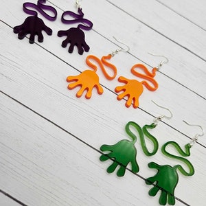 May include: Three pairs of hand-shaped earrings. The earrings are made of acrylic and come in purple, orange, and green.