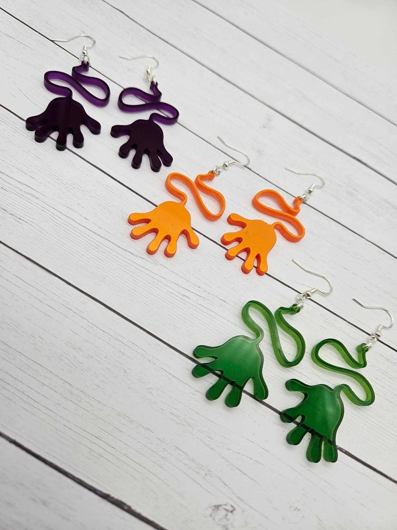 Peut inclure: Trois paires de boucles d'oreilles en forme de main. Les boucles d'oreilles sont en acrylique et sont disponibles en violet, orange et vert.