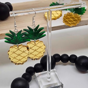 May include: A pair of gold pineapple earrings with green leaves on a clear acrylic earring stand. The earrings are on a wooden surface with black beads.