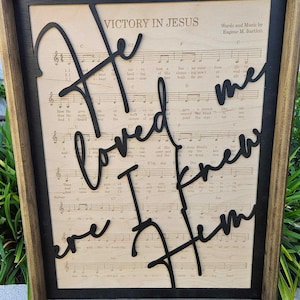 May include: A framed piece of sheet music with the words "Victory in Jesus" at the top. The words "He loved me, I knew Him" are cut out of black paper and layered on top of the music.
