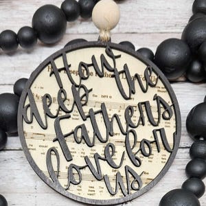 Könnte beinhalten: Runder Holzschmuck mit dem Schriftzug "How Deep the Father's Love For Us" in dunkler Farbe ausgeschnitten. Der Hintergrund zeigt Notenblätter. Der Schmuck hängt an einer Schnur und ist von schwarzen Holzperlen umgeben.
