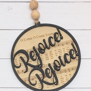May include: Circular wooden Christmas decoration with the word "Rejoice!" in a cursive font. The ornament has a black border and a background of sheet music. A jute string and wooden beads are attached for hanging.